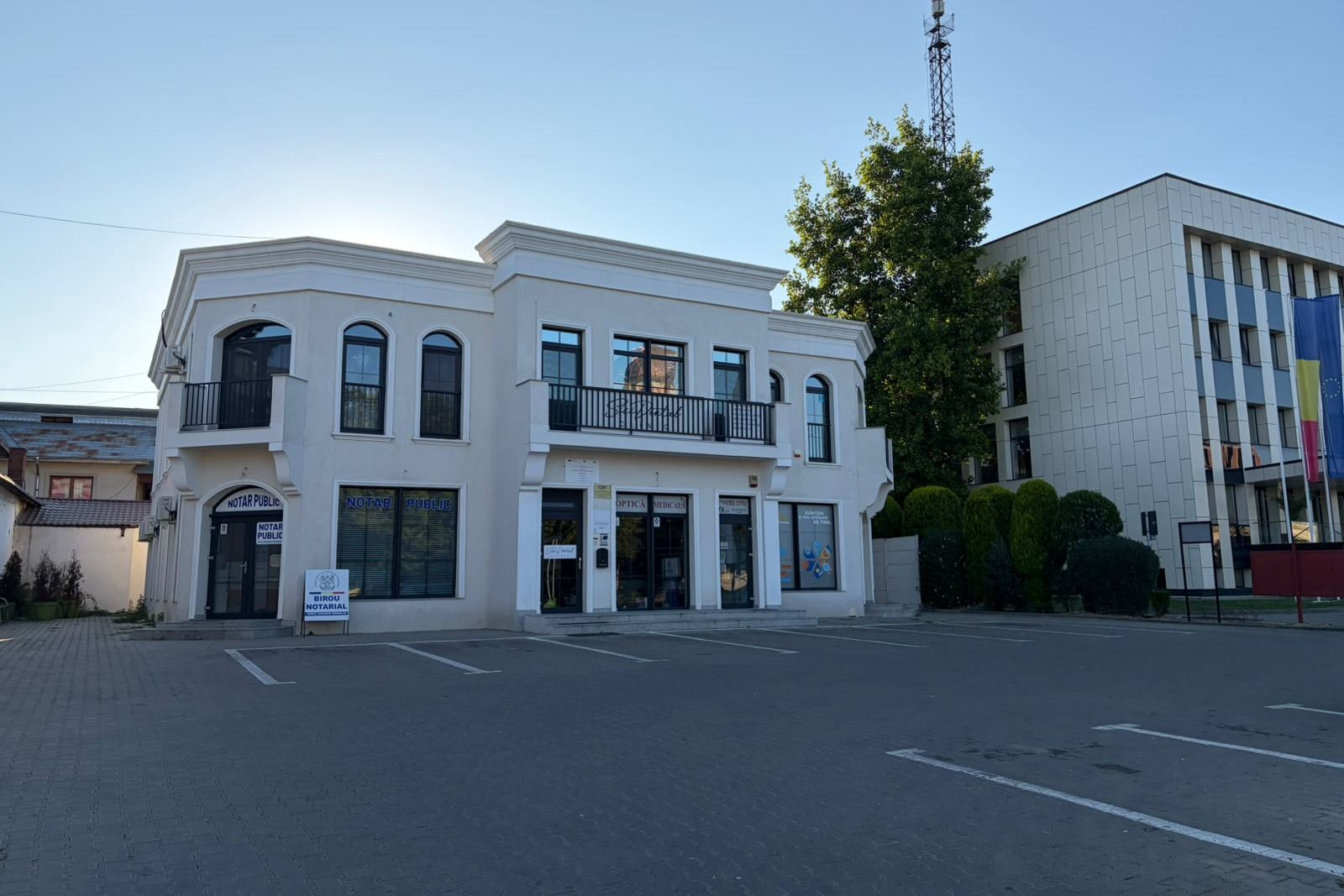 Clădirea albă în care funcționează biroul notarial, văzută dinspre parcarea din incintă, cu firmele „Notar public” la parter.